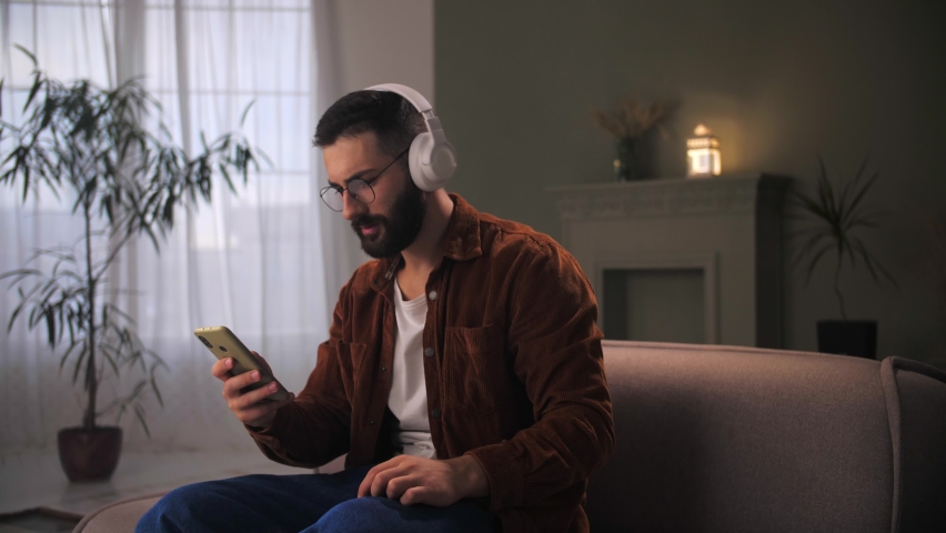 Young man celebration success using smartphone siting at couch.Bearded guy wear headphones looking at phone reading great news.Male winning lottery online cheering positive emotion. . High quality 4k | Shutterstock HD Video #1094690285