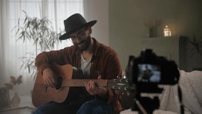 Male guitarist play song record video lesson online looking at camera.Man playing acoustic guitar sitting at sofa in home interior.Young bearded guy practicing play chords on instrument. High quality
