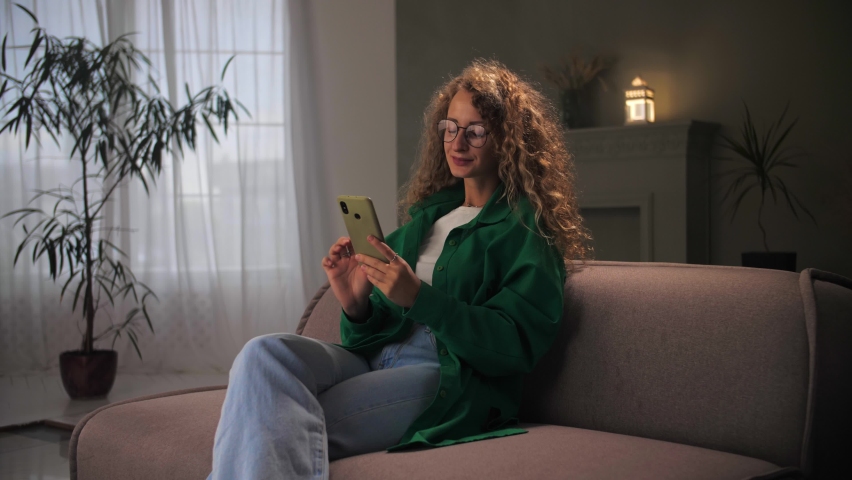 Woman browsing internet using phone sitting at couch.Young curly hair girl looking at smartphone smile and surfing web in home interior.Female wear eyeglasses holding cellphone doing online shopping | Shutterstock HD Video #1094690329
