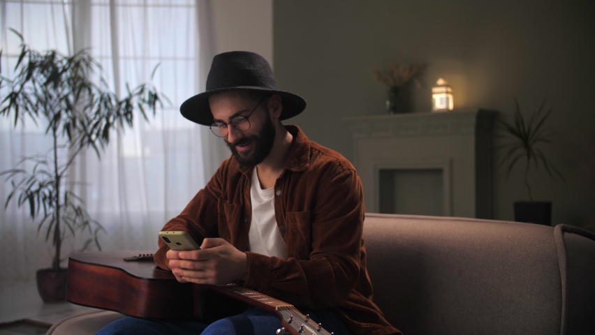 Young man celebration success using smartphone siting at couch.Bearded guy wear hat with guitar looking at phone reading great news.Male finished successful online study. High quality 4k footage | Shutterstock HD Video #1094690331