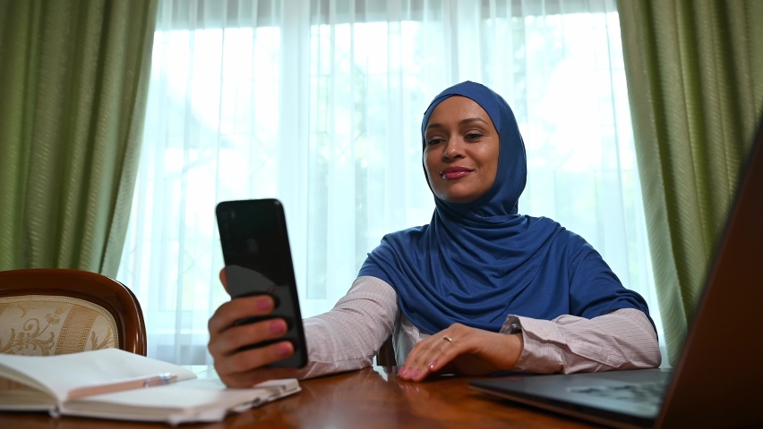 Muslim Middle Eastern pretty woman with head covered in hijab using smartphone, waves to her colleagues and business partners while communicates by video link. Video conference, call. Online meeting | Shutterstock HD Video #1094690903