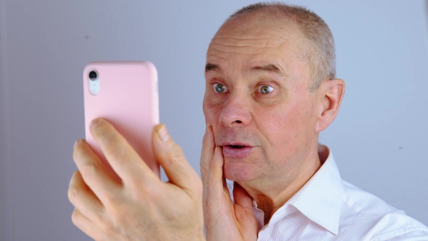 Close-up of mature charismatic man looks at screen of phone in pink cover, senior 60 years surprised, shocked by news, event, bright facial expressions, overhang, concept human stress resistance | Shutterstock HD Video #1094693465
