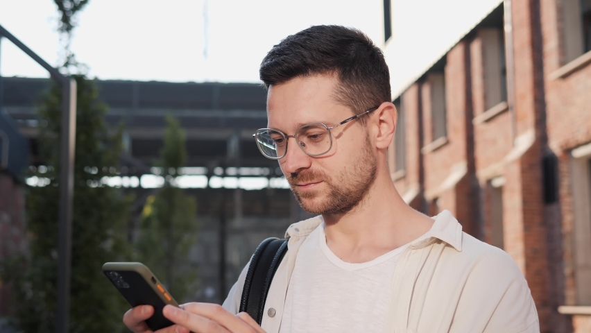 Young man in glasses winner looking at smartphone celebrate good news victory success standing outdoors at urban city background Excited overjoyed male winner celebrating success mobile phone victory. | Shutterstock HD Video #1094694049