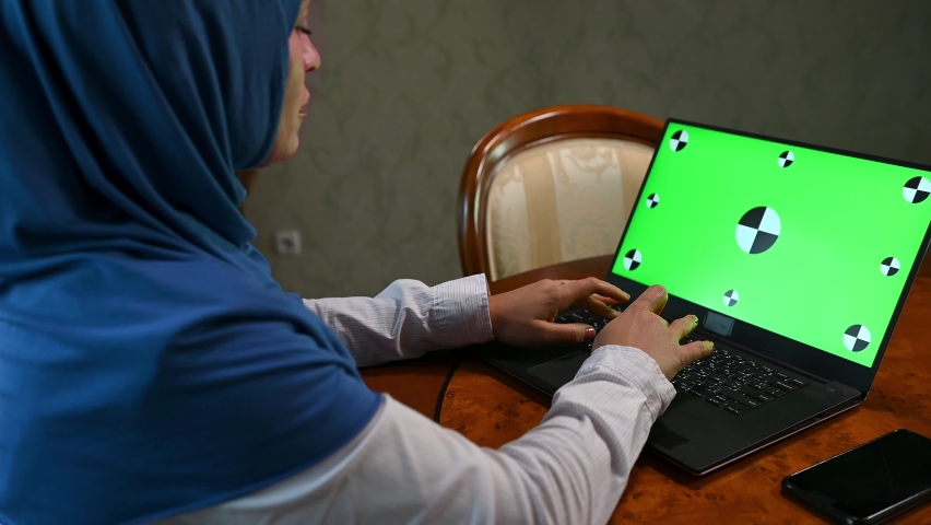 Rear view of a confident multitasking Arab Muslim woman in hijab, sitting at wooden desk and telecommuting, typing text on laptop with green chroma key mockup screen for insert promotional clip, video | Shutterstock HD Video #1094694855