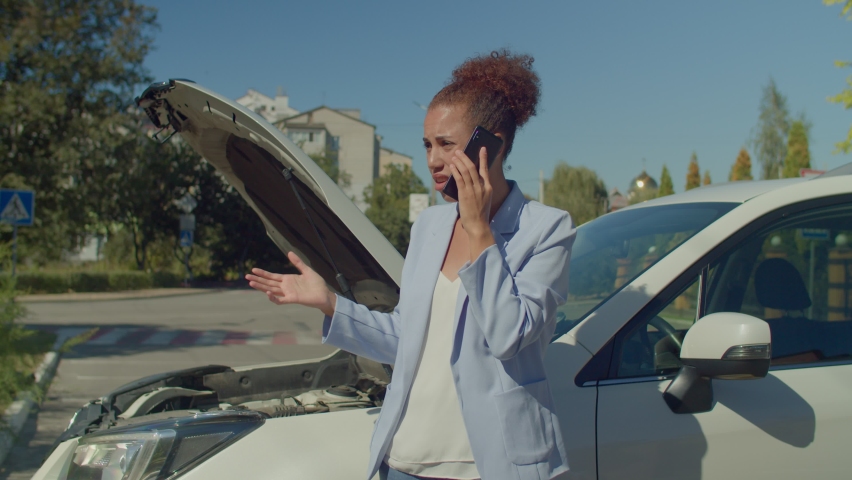 Upset beautiful black female driver standing near broken car with opened car hood, calling roadside assistance on cellphone while vehicle suffered mechanical failure during road trip in city. | Shutterstock HD Video #1094695057