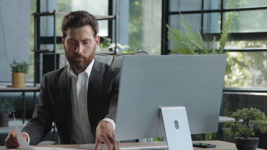 Caucasian pensive bearded man boss manager adult 40s businessman sitting in office busy with paperwork and computer check data in papers search information look at screen corporate company app project | Shutterstock HD Video #1094696011