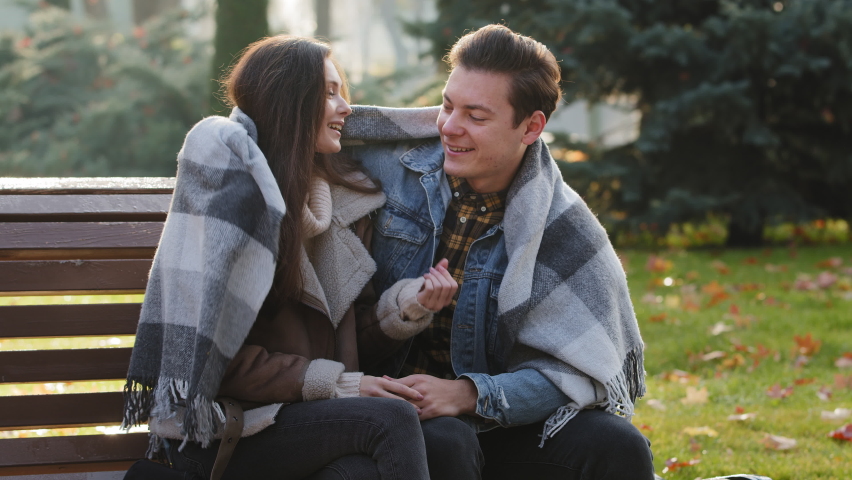 Cute loving couple sit on bench warming each other covering wool blanket outdoors in cold froze autumnal weather feel care love hugging funny talking with steam from mouth enjoy nature romantic date | Shutterstock HD Video #1094696127