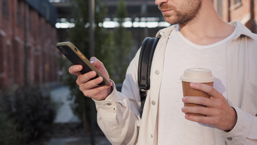 Stylish Man holding smartphone, chatting in social media outdoor. Good looking male with coffee to go messaging on mobile phone on urban street. Businessman typing a message outside, sunny morning. | Shutterstock HD Video #1094696165