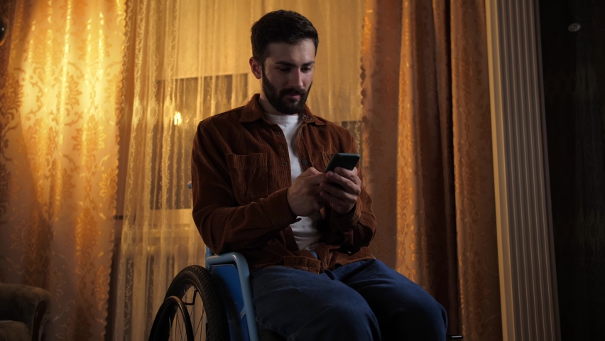 Young man in a wheelchair looking at phone and screaming indoors. Disabled guy Losing lottery using smartphone with sad mood in home interior.Male celebration victory concept. | Shutterstock HD Video #1094697515