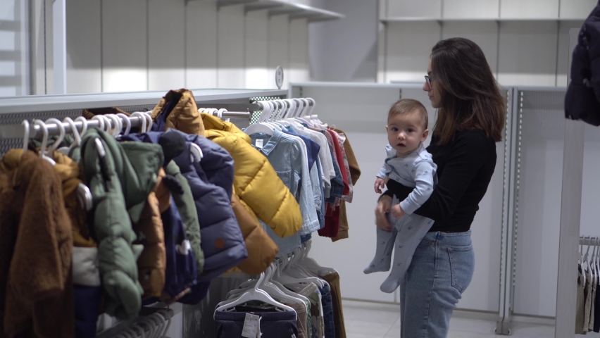 A young girl with a child in a boutique in the department with children's clothing. Mom and son in the store buys shirts for the child. | Shutterstock HD Video #1094704701