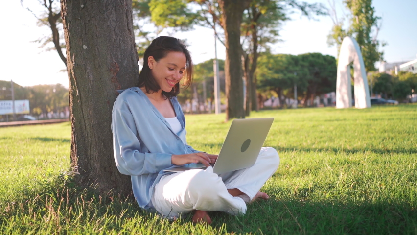 Happy brunette woman using laptop computer while sitting near the tree on grass in park. Young female freelancer working outdoors in city park. Caucasian businesswoman at remote work. Business person | Shutterstock HD Video #1094707285
