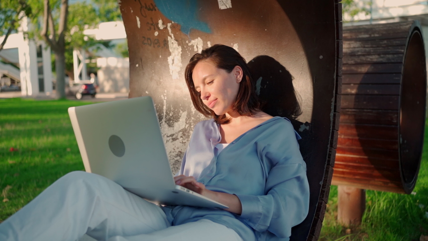 Happy brunette woman using laptop computer while sitting near the tree on grass in park. Young european woman sitting in park use laptop. Remote learning new normal | Shutterstock HD Video #1094707291