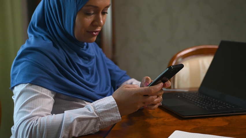 Beautiful pensive and serene young Arab Muslim woman with hijab, using mobile phone, typing a text, browsing social network, dreamily reading news, leaning on a wooden table at a cozy home interior | Shutterstock HD Video #1094708689