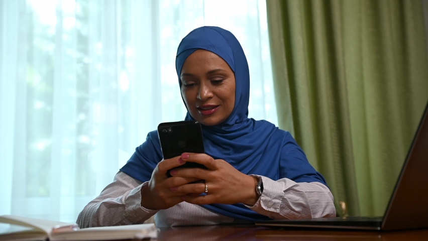 Charming confident stylish middle-aged Arab Muslim woman in hijab and formal wear, using mobile phone, scrolling news feed, browsing websites, checking content in social media, writing messages | Shutterstock HD Video #1094708709