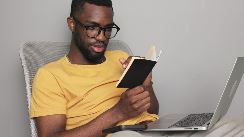 African american man in glasses looking in a notebook student and working in a laptop checking his knowledge at home online quarantine online learning | Shutterstock HD Video #1094716397