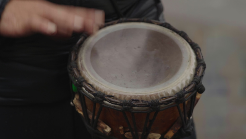 Close-up, the hands of a street musician beat the rhythm across the membrane of an African drum during a light rain near the roadway.