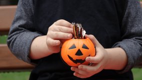 small cute halloween pumpkin bucket in kid little boy hand.child is smiling funny spooky.orange color bucket filled candies sweets wrapped in foil.kid hand take.park bench autumn fall trick or treat - Powered by Shutterstock - Get 15% off with code: PIKWIZARD15