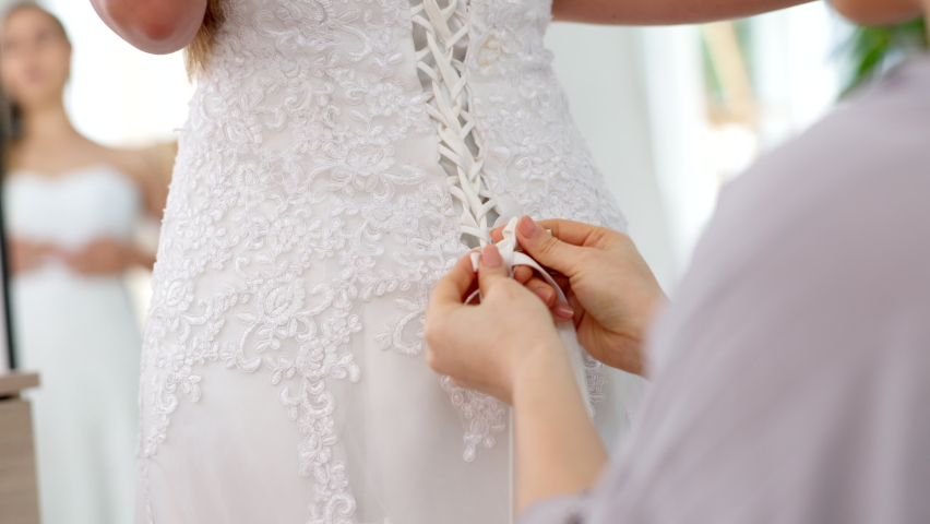 Friends helping woman with wedding dress knot in a room or boutique for marriage ready, fitting or fashion with corset lace detail and hands. Bride with mirror reflection getting ready for love event | Shutterstock HD Video #1094729989