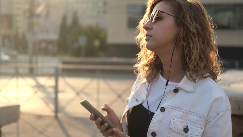 Attractive young woman in sunglasses listens to music on headphones, uses a smartphone on a walk in the city at sunset. Close-up portrait of a girl who sings and dances | Shutterstock HD Video #1094731027
