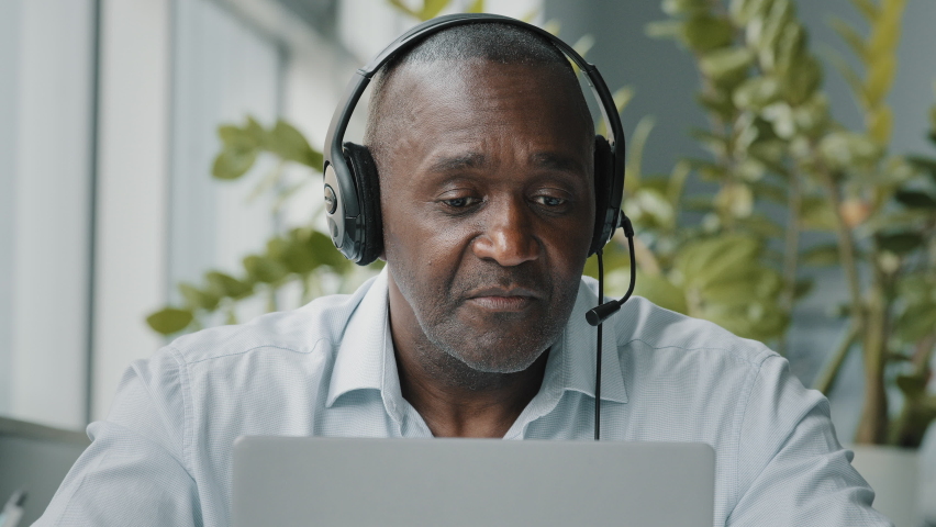 Senior african employee sit at office desk wear headphones with microphone connect to video conference business call on laptop communicate by webcam talk in online chat does thumb up approval gesture | Shutterstock HD Video #1094733603