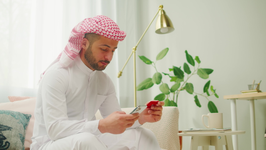 Middle eastern man using credit card and smartphone, shopping in app. Male person buying clothes in application, online order, food delivery. Wearing traditional Islamic clothes, relaxing at home. | Shutterstock HD Video #1094737099