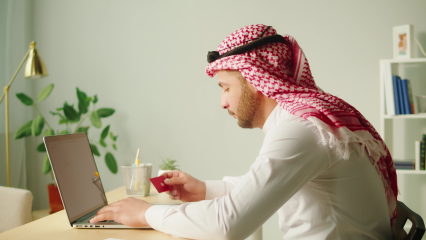 Middle eastern man using laptop. Home office. Male person texting at computer keyboard in living room. Wearing traditional Islamic clothes. Communicating with family and friends online. | Shutterstock HD Video #1094737127