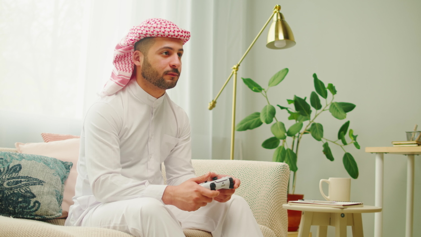Middle eastern man playing video games on modern console. Male person using white gamepad, enjoying AR 3D game in living room. Wearing traditional Islamic clothes, relaxing at home. | Shutterstock HD Video #1094737133