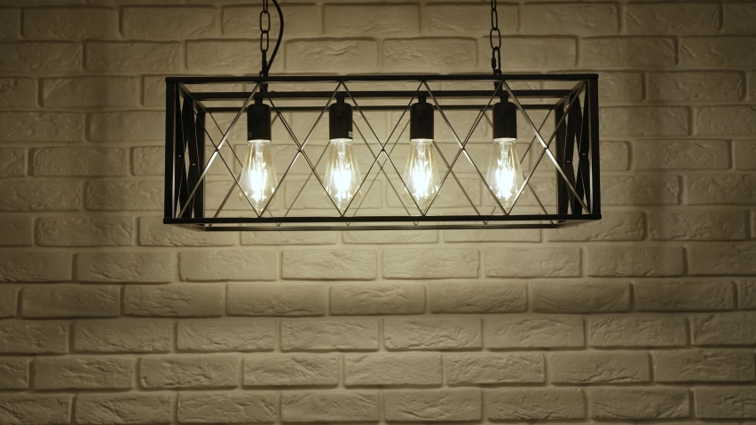 Four filament lamps in pendant lamp turn on and off, close-up. Lamps in black metal box are hung on chains near white brick wall in a loft apartment. | Shutterstock HD Video #1094747171