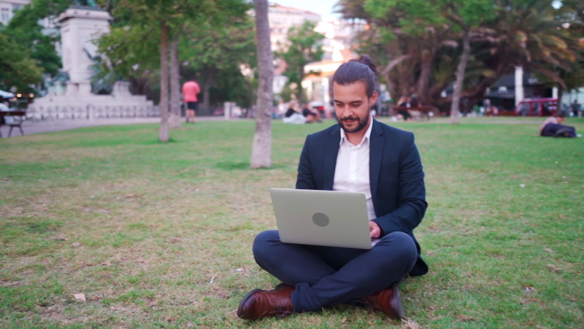 Handsome hispanic male businessman sitting lotus position in public park use laptop. Remote work concept. Stylish freelancer in suit relax in park summer day working on computer. Technology. Handheld. | Shutterstock HD Video #1094748349