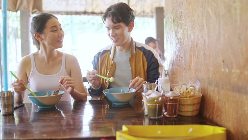 Happy Asian couple enjoy outdoor lifestyle in the city shopping and having lunch eating street food noodle together at chatuchak weekend street market in Bangkok, Thailand on summer holiday vacation.