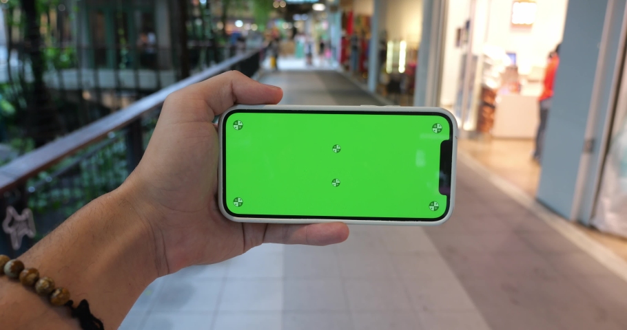A man's hand holds a mobile phone horizontally with copy space to insert a promotional presentation, video clip or advertising text, on green chroma key screen, standing in a megapolis shopping mall | Shutterstock HD Video #1094752857