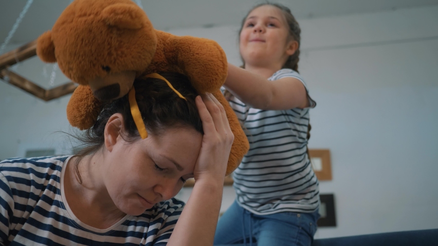 Irritated girl with sore head. Daughter screams and plays with teddy bear. Loud screams annoy sick mother in quarantine. Screaming of children disturbs parent. Mom with sore head is in quarantine. Royalty-Free Stock Footage #1094757681