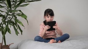 Cute little kid girl sitting on bed holding smart phone enjoy using digital tablet, playing games at home. Small child learning in tablet, watching video. Mobile technology concept. 