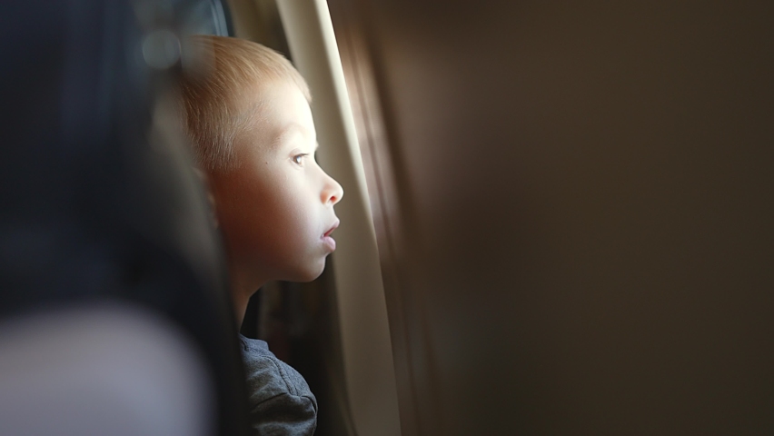 Side view from back seat, child looks out window of an airplane in flight. Tourist travel lifestyle. Looking aerial view of sky and cloud. Look around indoors. Sleepy tired baby boy in transport. Royalty-Free Stock Footage #1094765025