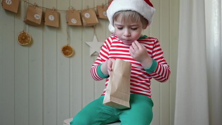 Boy in pajamas, Santa cap eating sweet candy on paper advent calendar with presents background. Little happy child with gift of handmade Christmas calendar on wall. Celebrating at home Xmas tradition. | Shutterstock HD Video #1094766283