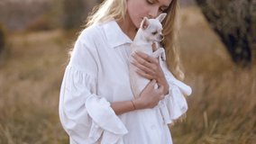 Close-up video of an adorable long-haired blonde girl holding a shaking white chihuahua. A woman loves her dog, caresses it. little friend A pet in autumn nature