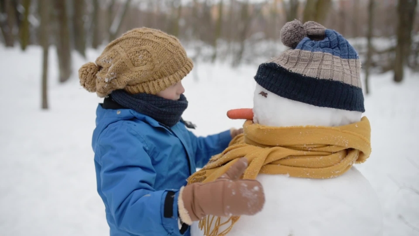 Little boy building snowman in snowy park. Child puts hat and scarf on snowman . Active outdoors leisure with family with children in winter. Kid during stroll in a snowy winter park Royalty-Free Stock Footage #1094768369