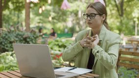 the young student studies outdoors. Young woman with eyeglasses, sits at a cafe in the park in front of a computer. He stretches his hands in front of him, tired from hitting the keys on the computer.