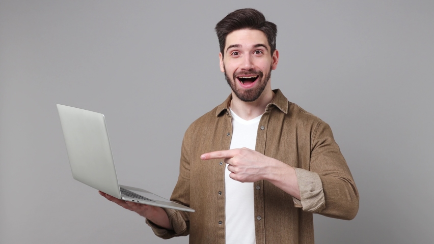 Young happy caucasian IT man 20s he wear brown shirt hold in hands typing use work point index finger on laptop pc computer isolated on plain grey background studio portrait. People lifestyle concept | Shutterstock HD Video #1094772733