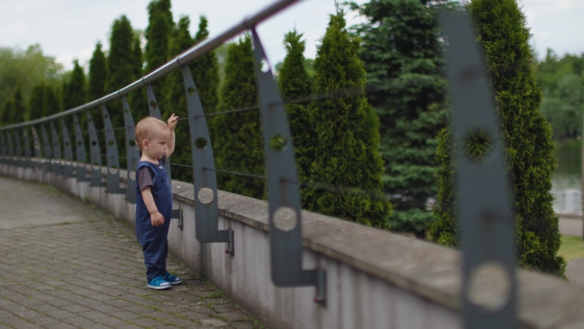 lonely baby child holding metal barrier string of fence looking behind fencing. concept of ban prohibition, limits and restrictions. poverty lifestyle. male kid child walking alone outdoors in daytime