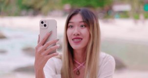 Millennial Asian woman talking looking at mobile phone camera sitting close the beach