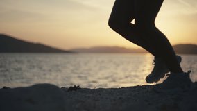 Women's legs in sneakers run on the rocks by the ocean at sunset. Close-up of athletic legs jogging at sunset. Slow motion video.