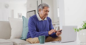 Video of senior biracial man using smartphone and laptop sitting in living room, smiling. Retirement, communication, inclusivity and senior lifestyle concept.