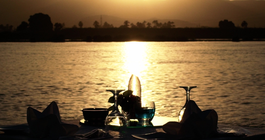 Golden hour shot of a romantic table set alongside a river, the table is set for two persons only with a pair of tissues and a pair of glasses on the table, sun reflection makes river water seems gold