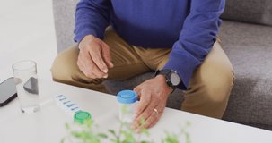 Video of midsection of senior biracial man sitting and taking pills at home. Retirement, medicine, healthcare, inclusivity and senior lifestyle concept.