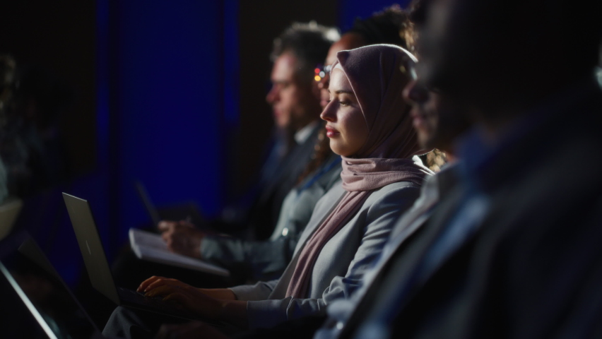 Arab Female Sitting in a Dark Crowded Auditorium at a Human Rights Conference. Young Muslim Woman Using Laptop Computer. Activist in Hijab Listening to Inspiring Speech About Global Initiative.