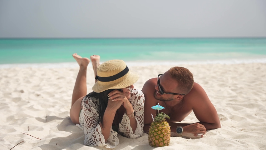 Latin couple lies and talking on the white sand on the tropical beach. Pineapple cocktail and paradise pleasure against the background of the turquoise sea. Slow motion shot Royalty-Free Stock Footage #1094804405