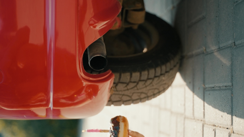 Vertical video. Comedy funny video. Car birthday celebration. Smoke from exhaust pipe of car blows out flame of candle. Candle in donut with chocolate. Close-up of girl