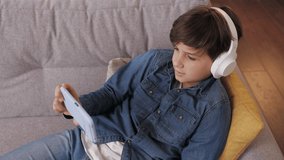 Young cute boy holding smartphone sitting at home. Teenager wearing earphones choosing favorite music, texting messages, browsing internet, watching video in social media, playing online games.