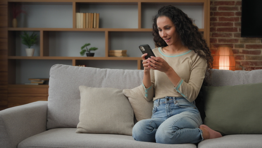 30s Hispanic Latina Caucasian girl woman lady sitting on couch at home living room play with mobile phone win game online betting victory has discount in net virtual hop e-commerce triumph celebrate Royalty-Free Stock Footage #1094807531